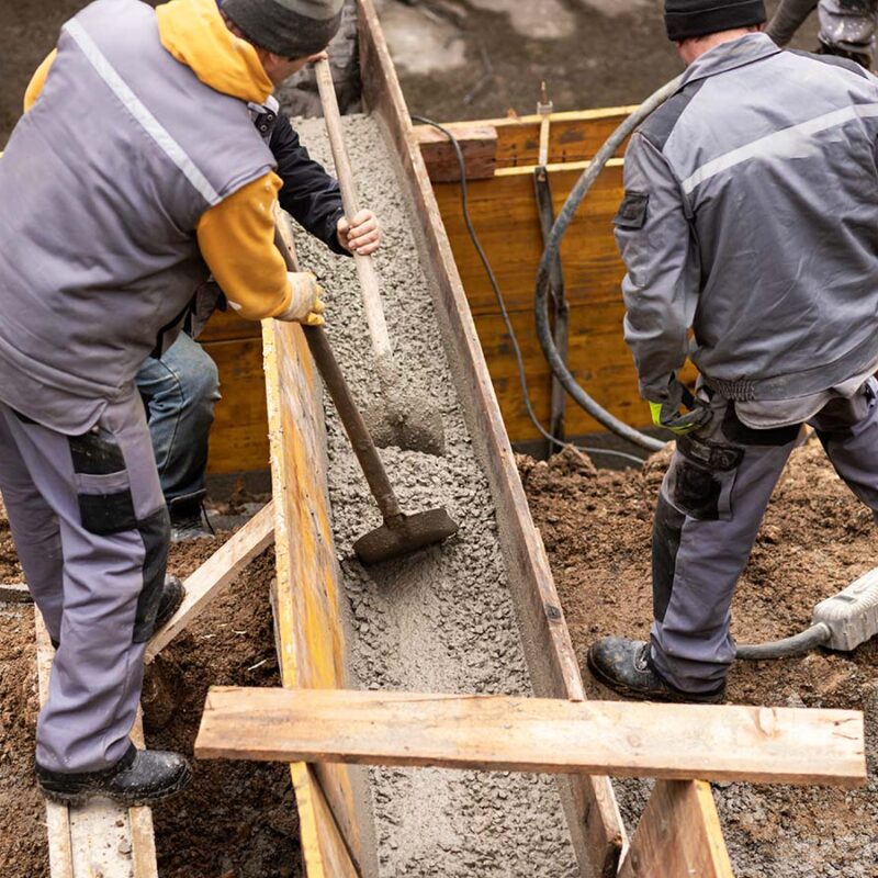 A man wearing a yellow hard hat is pouring cement into a bowl