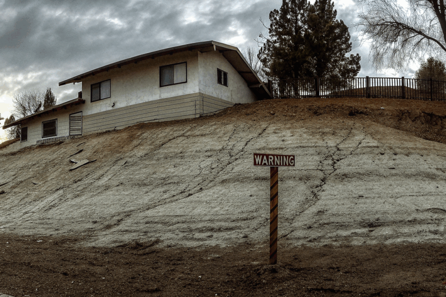 A two-story house with visible foundation cracks and a broken driveway due to hillside soil creep, with a leaning fence and dry slope in the background.